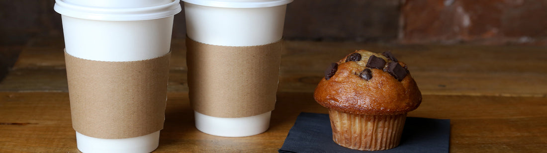 white coffee cups in kraft cup sleeves beside a chocolate chunk muffin on a black napkin