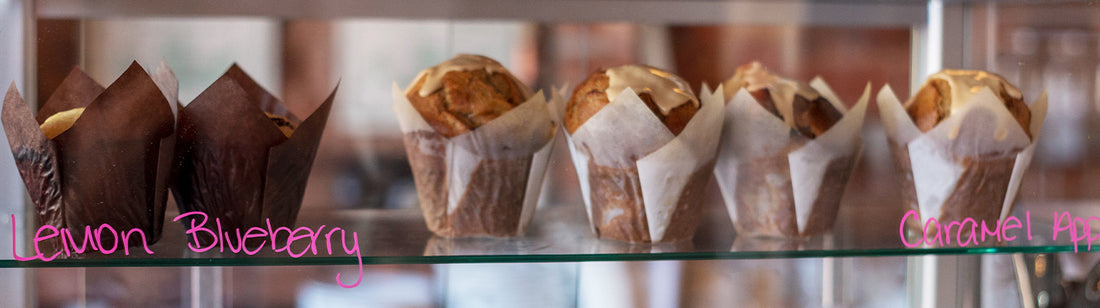 A glass display with lemon blueberry muffins in brown fluted baking cups and caramel apple muffins in white fluted baking cups