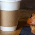 two white coffee cups with cup sleeves next to a chocolate chunk muffin on a black napkin