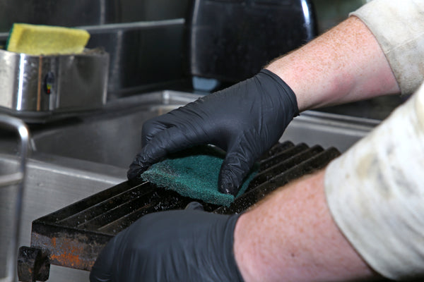 person wearing black nitrile gloves and scrubbing grill with Green 6